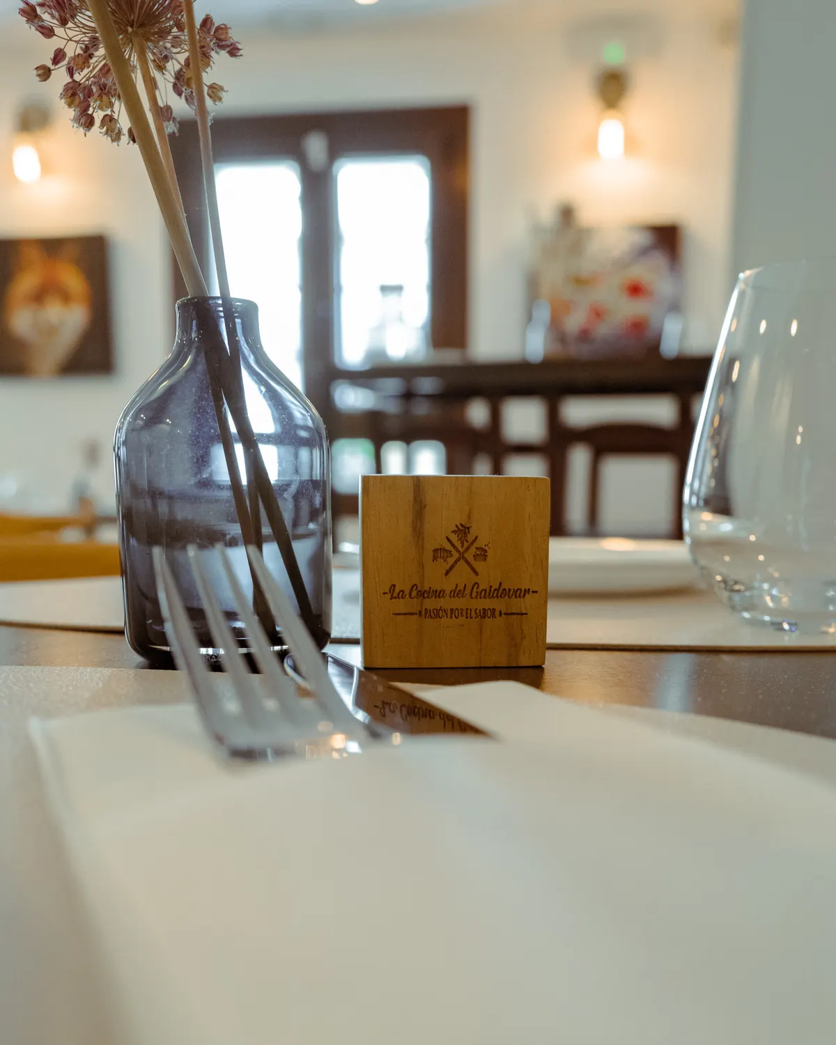 Mesa del comedor de La Cocina del Gaidovar con un taco de madera grabado con su logo, una copa y un jarrón de flores secas